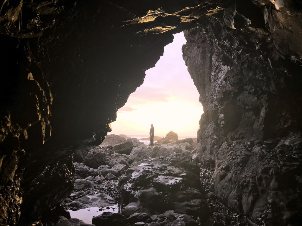 Looking out of a small sea cave at a man standing on rocks in the distance, surveying the scene during a pretty sunset on the Oregon Coast.