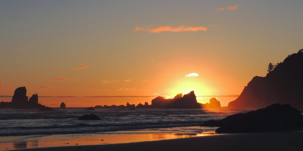 The sun passes behind a big offshore rock during an orange sunset at Crescent Beach in Ecola State Park, Oregon.