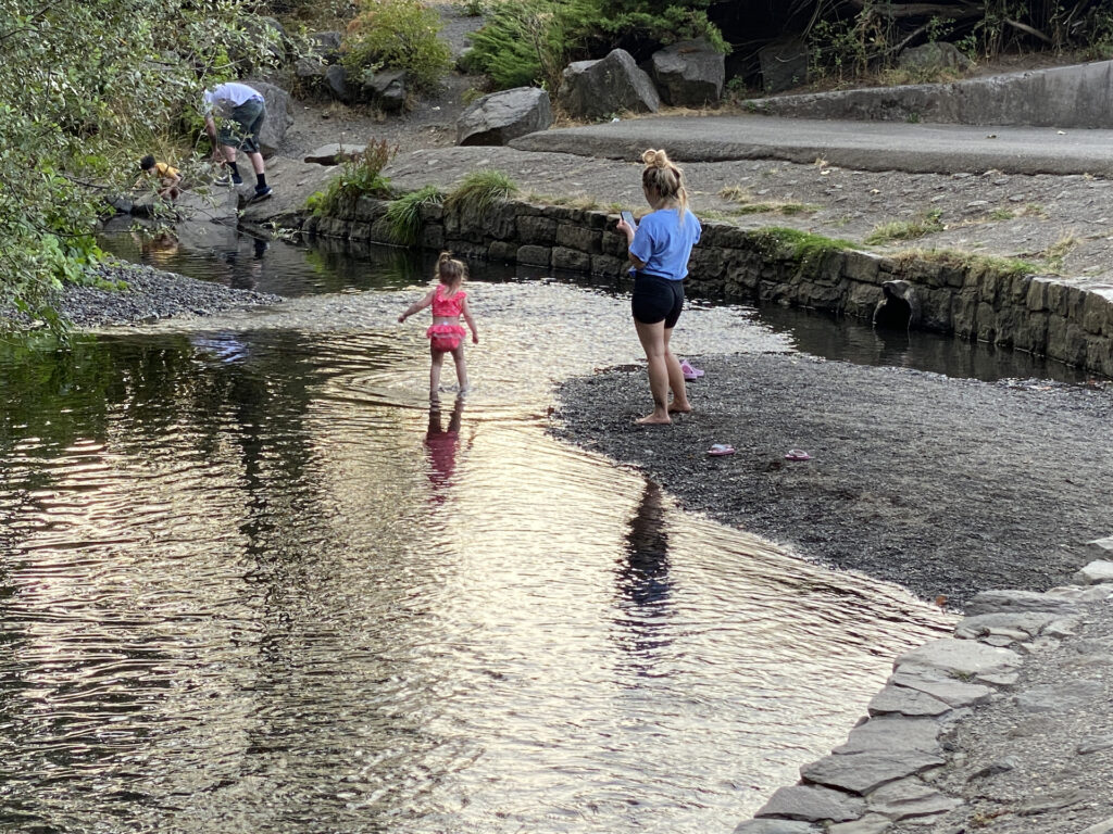 A toddler in a pink swimsuit wades in Multnomah Creek as mom watches nearby. 