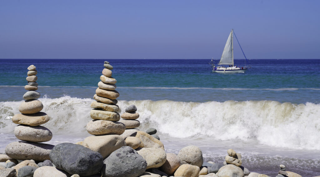 Three stacks of smooth stones stand on a rocky shore as a wave breaks behind them, with a sailboat gliding by in the distance over blue ocean water.