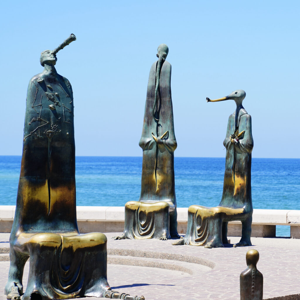 Three metal chairs with very tall backs topped with fanciful figures sit empty along the Malecon in Puerto Vallarta. Behind them is the vast blue Pacific Ocean.