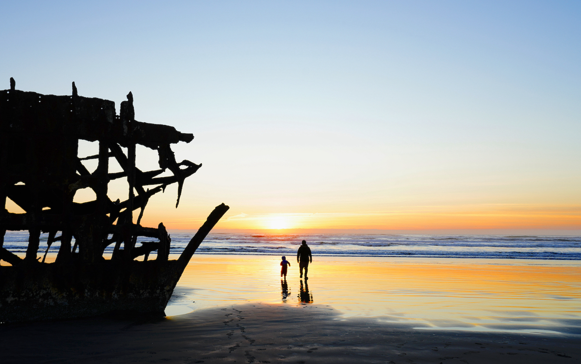 A toddler and his dad walk towards the ocean sunset next to the Peter Iredale shipwreck at Fort Stevens State Park on the Oregon Coast.