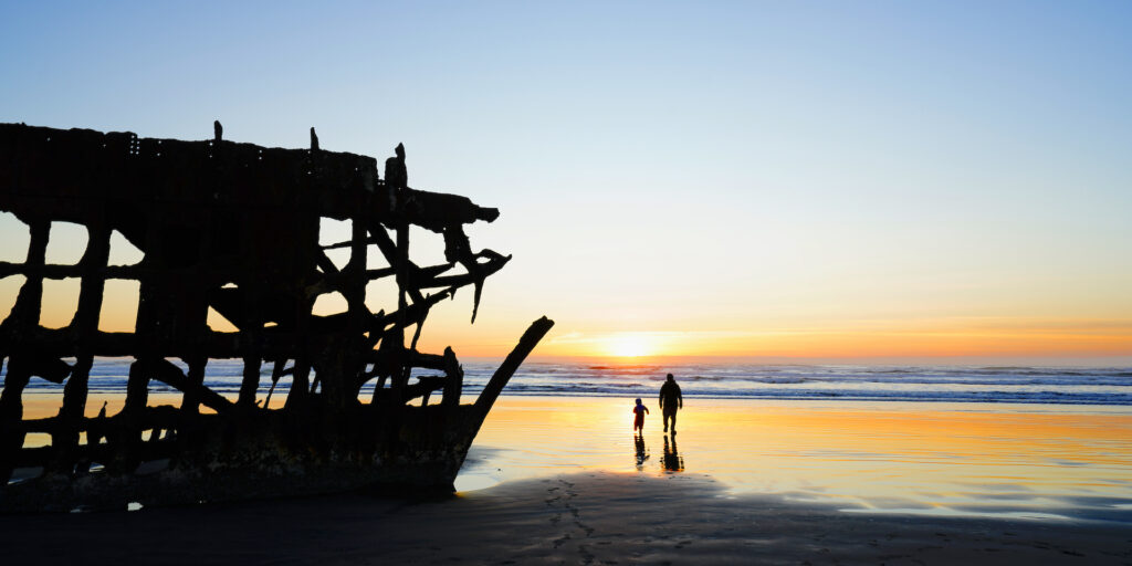 A toddler and his dad walk towards the ocean sunset next to the Peter Iredale shipwreck at Fort Stevens State Park on the Oregon Coast.