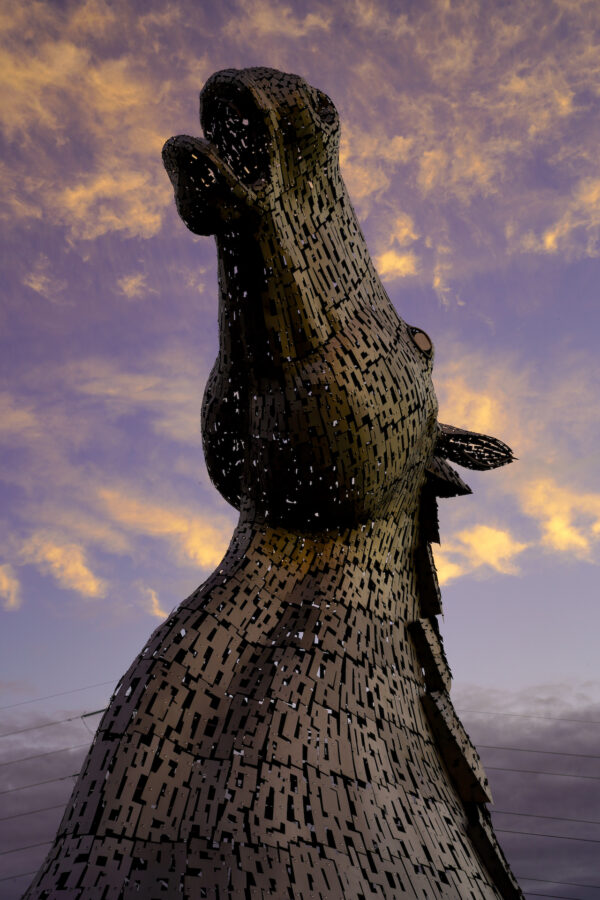 A massive modern metal sculpture of a horse's head looking up stands against orange sunset clouds.