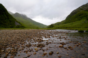 Looking up a shallow river and rocky shore toward the green slopes of Glen Nevis in Scotland.