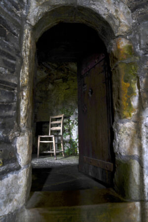 Looking through an arched stone doorway to a little room with one wooden chair inside.