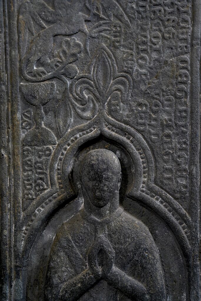 A detail of a carved stone grave marker at Iona Abbey shows a figure with hands folded in prayer.