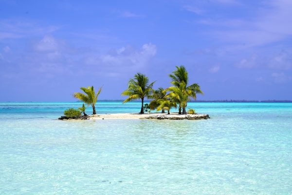 A small sand island with a few palm trees rises from the pale aquamarine water of the lagoon around Taha'a.