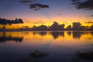 Bands of gold, pink, and periwinkle glow behind dark clouds, all reflected in the calm lagoon on Moorea.