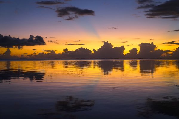 Bands of gold, pink, and periwinkle glow behind dark clouds, all reflected in the calm lagoon on Moorea.